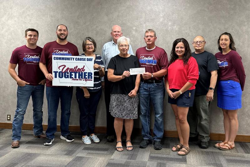 Nine people stand against a gray wall in an office setting. The two people on the far left are holding a holding a sign that reads "Community Cause Day: United Together: Making the Difference. In the center of the group, two people are holding a paper check.