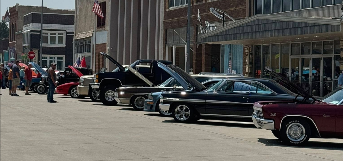 A view of several cars parked, facing the center of the road, with their hoods popped open.