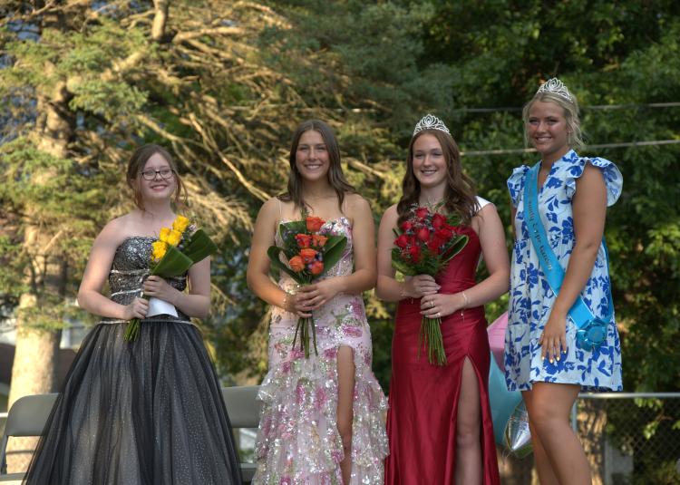 A far away shot of four women standing on stage, facing the camera. Riley Dobbins is wearing a black and silver strapless floor-length gown. Morgan McChesney's dress is pink with darker pink flowers and green leaves, also floor-length. Madison Becker's gown is red satin and ankle length. Katie Vogel is wearing a light blue dress with dark blue flowers that hits just above the knee.Calhoun County Phoenix - Staff Photo - Create Article