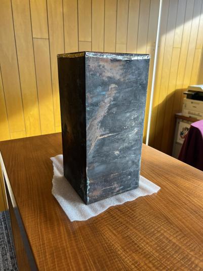 A close-up photo of a grey metal box sitting on a wooden desk. The box, a time capsule, is approximately 18 inches high and 11 inches by 11 inches square.
