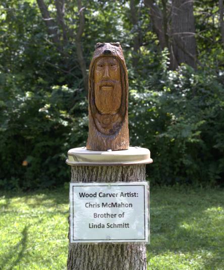A wooden carving depicting a bear's open mouth with a bearded man's head inside. A sign reads "Wood carver artist: Chris McMahon, Brother of Linda Schmitt"